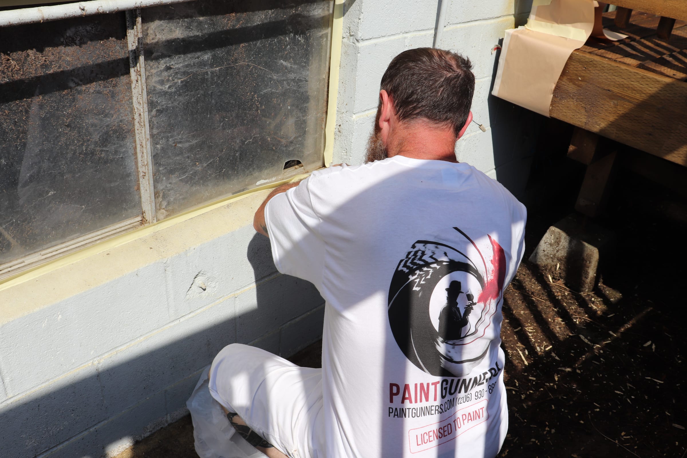 Painter kneeling to seal the bottom edge of a masked window before exterior painting
