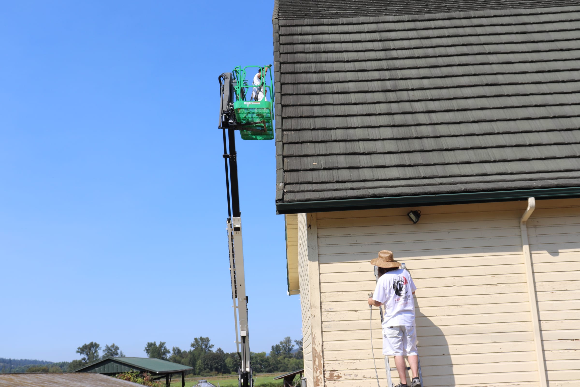 Boom lift extended along a tall barn wall while a second painter works at ground level