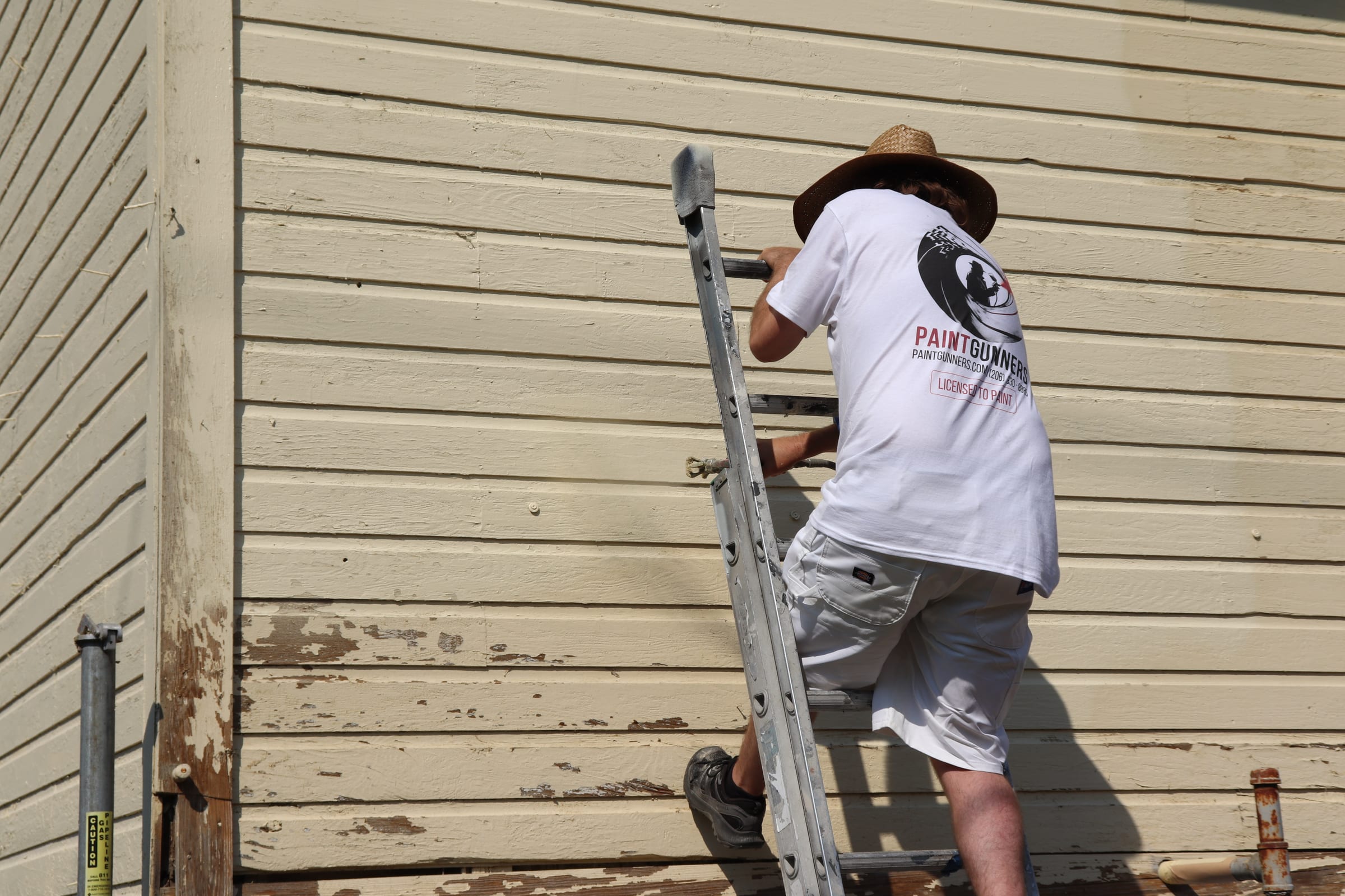 Painter spraying siding from a ladder with weathered bare boards visible below the fresh coat