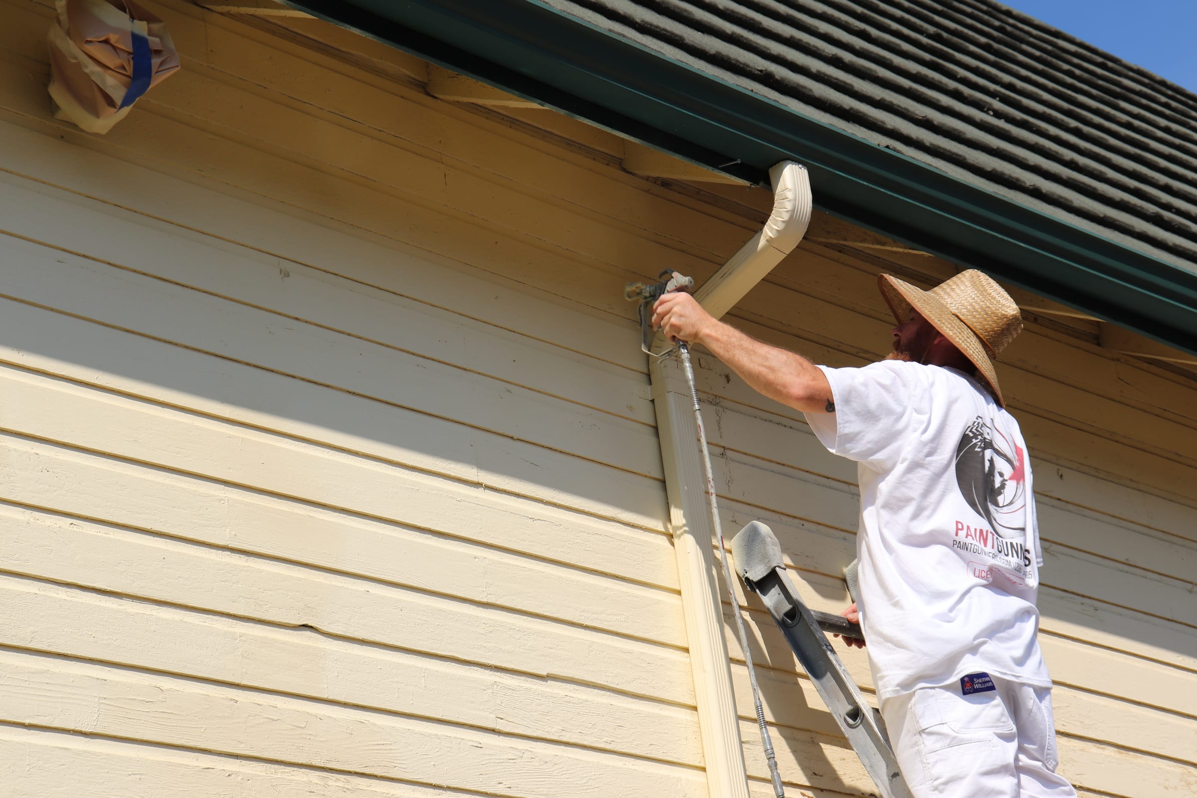 Painter coating a gutter and downspout alongside freshly painted siding