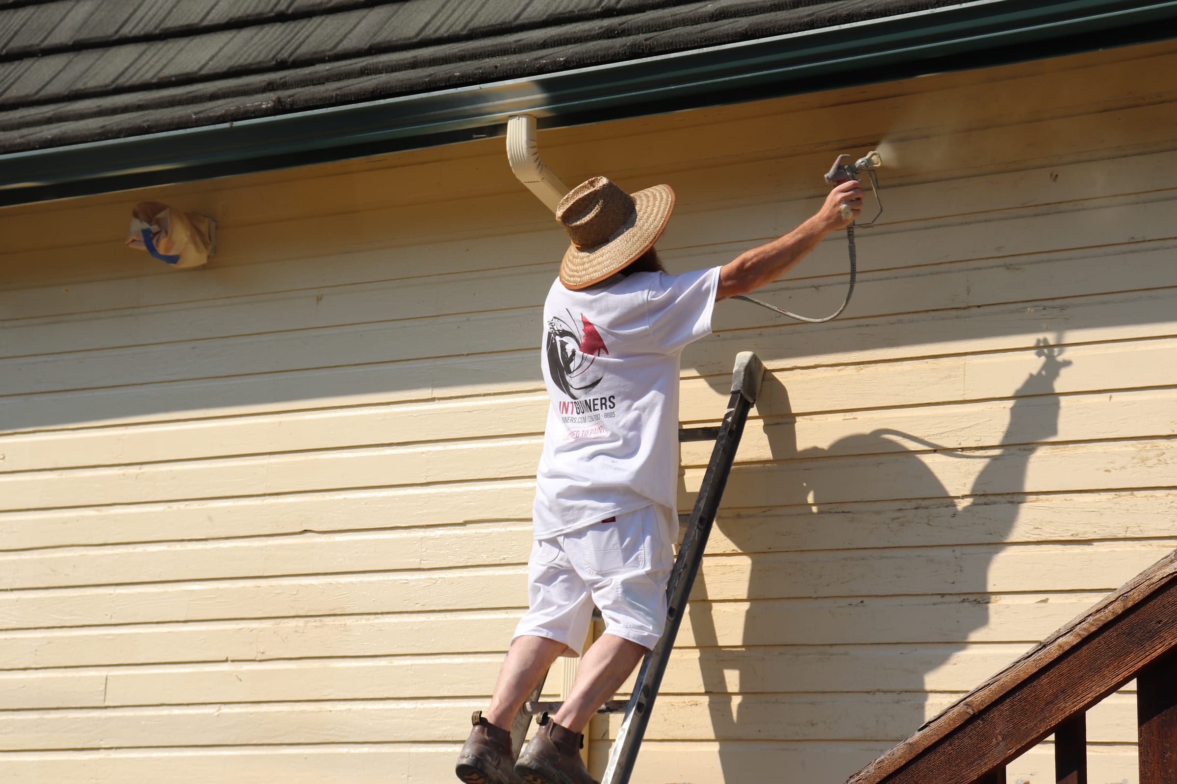 Painter on a ladder spraying paint along the eave line of a building