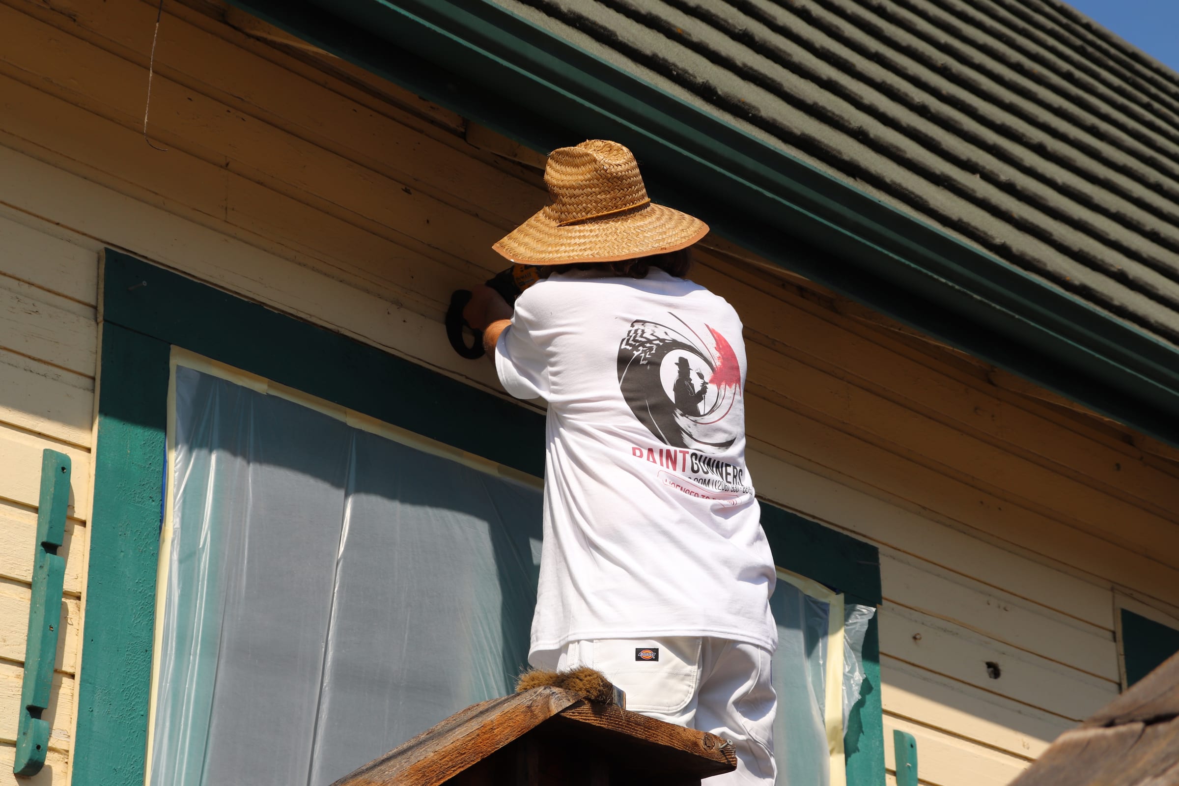Painter in a branded shirt working around a masked window on cream siding