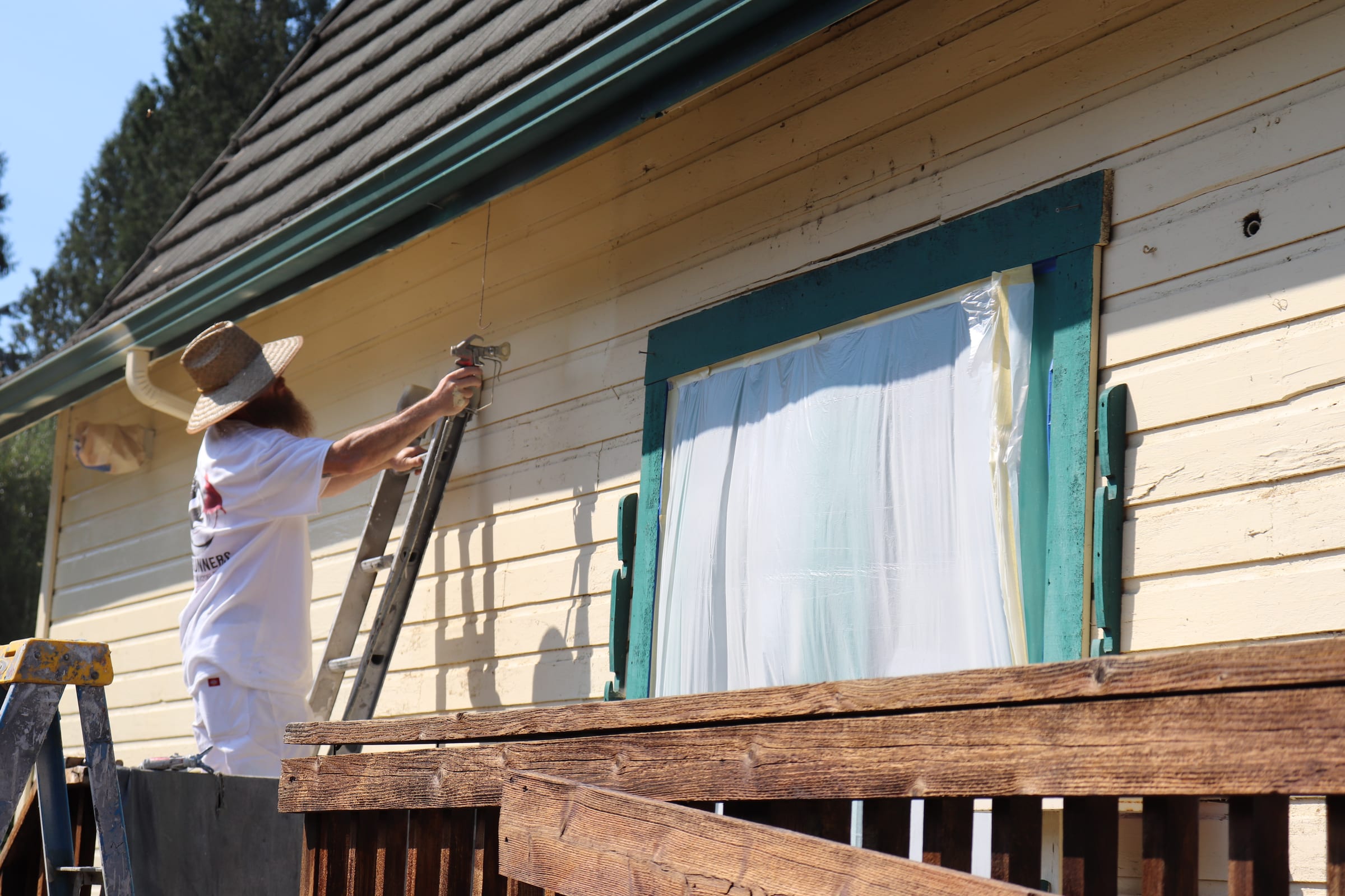 Painter spraying siding from a ladder beside a masked window bay