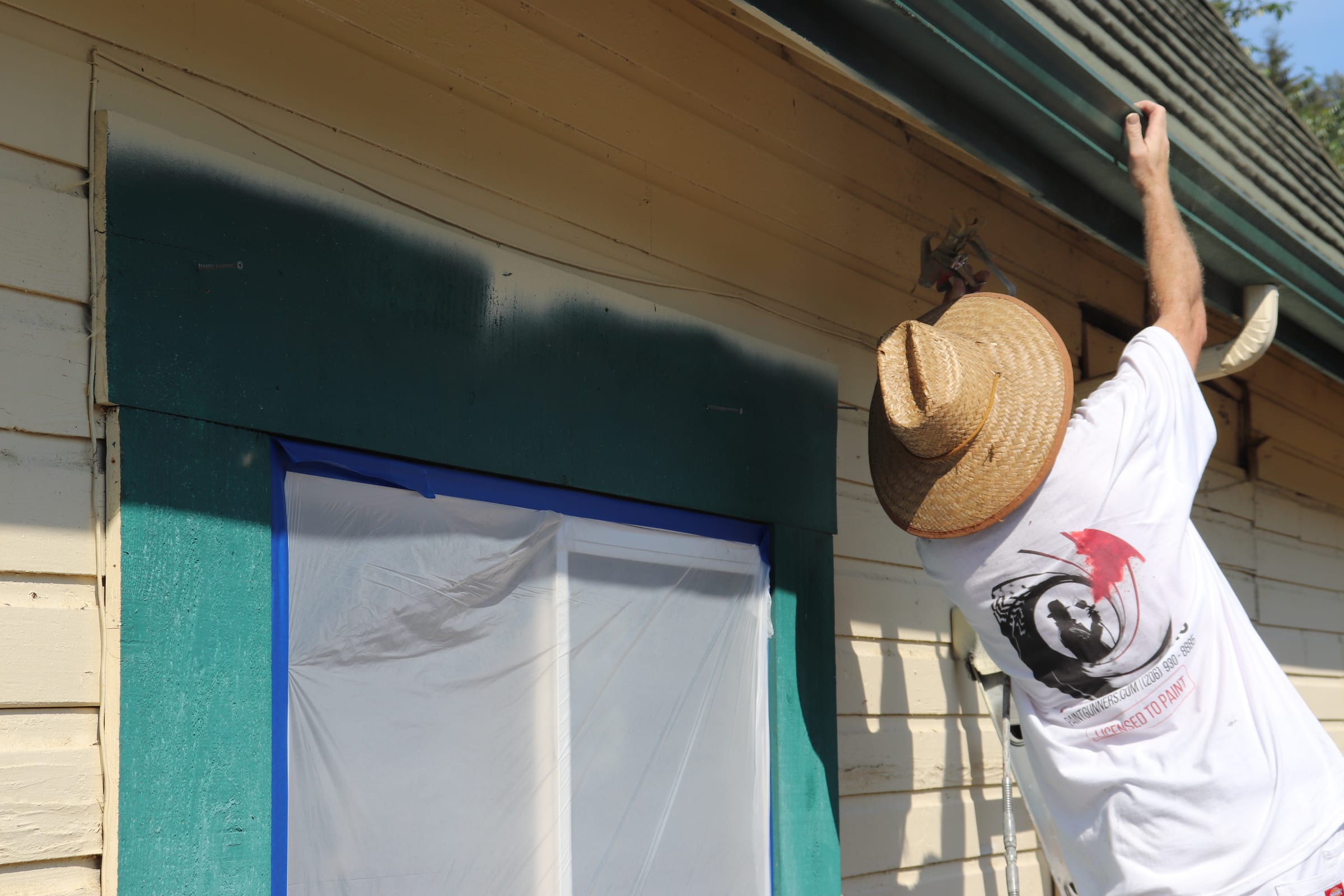 Painter working the eave above a window masked with plastic and blue tape