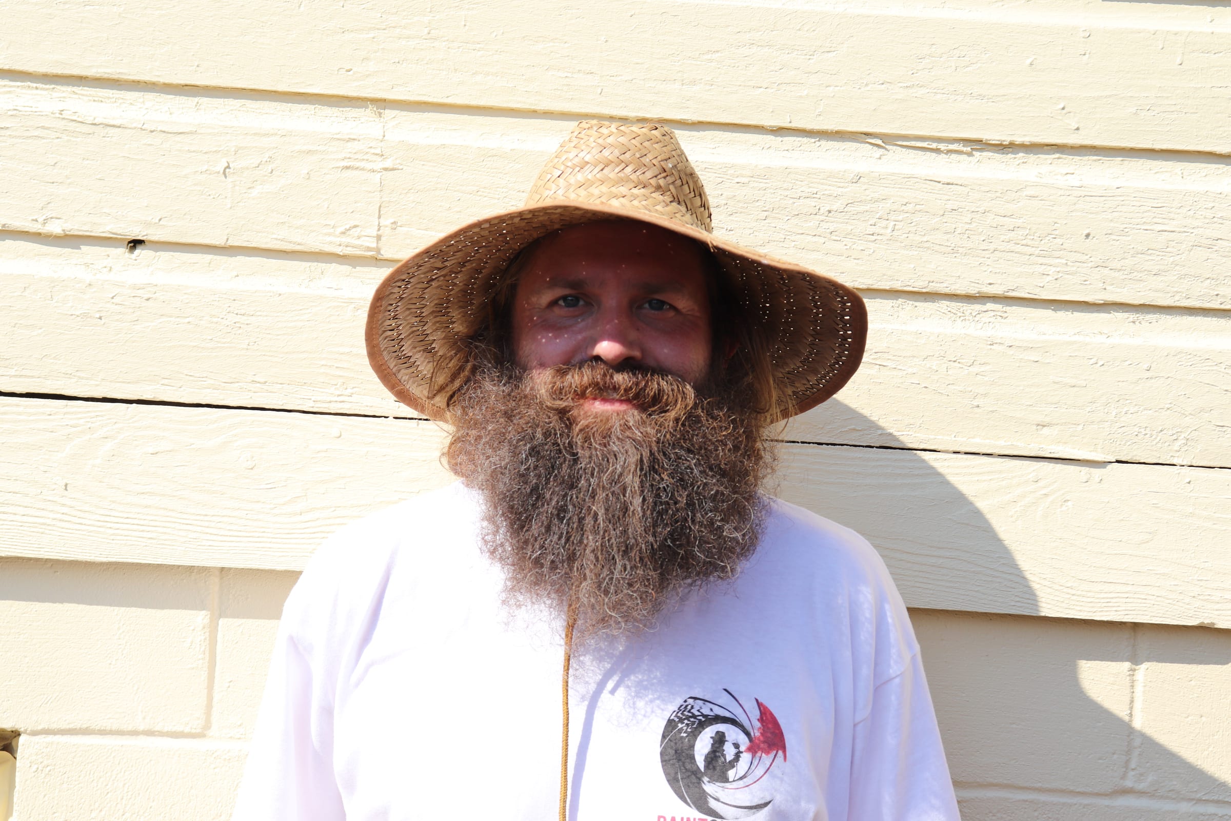 Bearded Paint Gunners painter in a straw hat in front of freshly painted siding